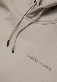Beige huppari, jossa on nyörit ja mustat päätylenkit. "PeakPerformance" -logo brodeerattu kontrastoivalla langalla rinnassa. Pehmeä pinta.