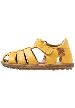 Sandalia de cuero amarillo con diseño abierto, presenta correas cruzadas y cierre de velcro. Suela de goma texturizada con pequeños patrones circulares, detalle del logo de la marca.