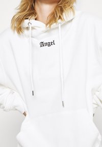 Bílá mikina s kapucí vyrobená z měkké látky, s černým výšivkou "Angel" na hrudi, šňůrkami s kovovými konci a volným střihem.