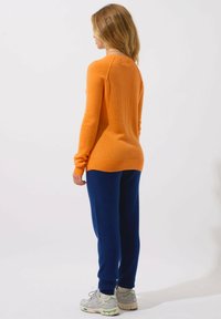 Maglione arancione lavorato a maglia con maniche lunghe, polsini a coste e scollo rotondo, abbinato a pantaloni blu a vestibilità rilassata e sneakers grigie.