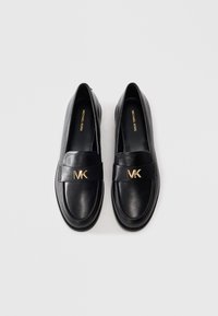 Een paar zwarte leren instappers van Michael Kors met een gouden MK-logo op de band, gezien van bovenaf op een effen lichte achtergrond.