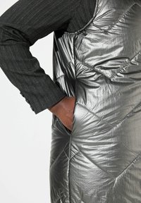 Silberne, gepolsterte Weste mit einem gesteppten Design, ausgestattet mit einer Seitentasche und einem schwarzen Hemd mit Fischgrätmuster darunter.