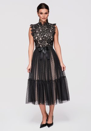 Donna che indossa un vestito nero in pizzo senza maniche con cintura a fiocco in raso e gonna di tulle trasparente a pois, abbinato a tacchi neri.