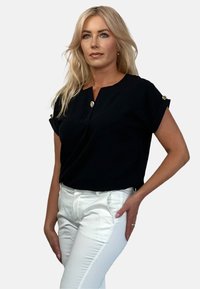 Czarna bluzka z krótkim rękawem i dekoltem w kształcie litery V, z złotymi guzikami na rękawach, w połączeniu z dopasowanymi białymi spodniami. Gładka tekstura materiału.
