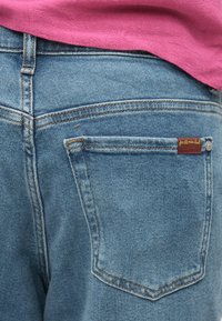 Buzunar din spate al pantalonilor din denim albastru deschis, cu o etichetă roșie și o persoană care poartă un top roz.