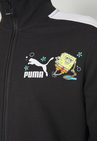 Veste noire avec logo Puma blanc et Bob l'Éponge tenant une épuisette à méduse, avec de petits motifs de fleurs bleues et de bulles.