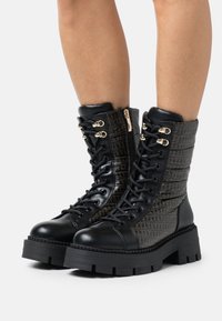 Bottines noires en cuir lisse et tissu à motifs, présentant des éléments dorés, un laçage à l'avant et des semelles en caoutchouc épaisses.