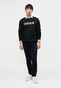 Μαύρο φούτερ με γραφικές εκτυπώσεις στο στήθος, συνδυασμένο με ανοιχτό γαλάζιο πουκάμισο με γιακά από κάτω, σκούρο τζιν και λευκά αθλητικά παπούτσια.