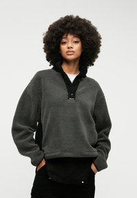 Πουλόβερ φλις σε σκούρο γκρι χρώμα με ψηλό γιακά και μαύρες λεπτομέρειες. Διαθέτει μαλακή υφή και μπροστινές τσέπες.