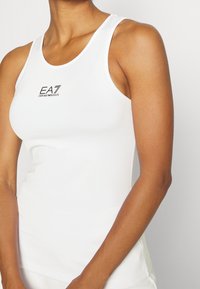 Kvinna klädd i en figursydd vit ärmlös topp med "EA7 Emporio Armani"-logotyp på bröstet, mot en enkel ljus bakgrund.