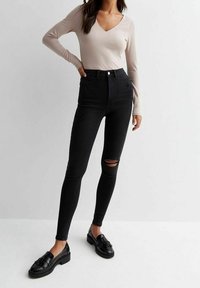 Jeans negros de cintura alta con corte ajustado, que presentan un desgaste en la rodilla, combinados con una blusa de manga larga en color beige claro con escote en V y mocasines negros.