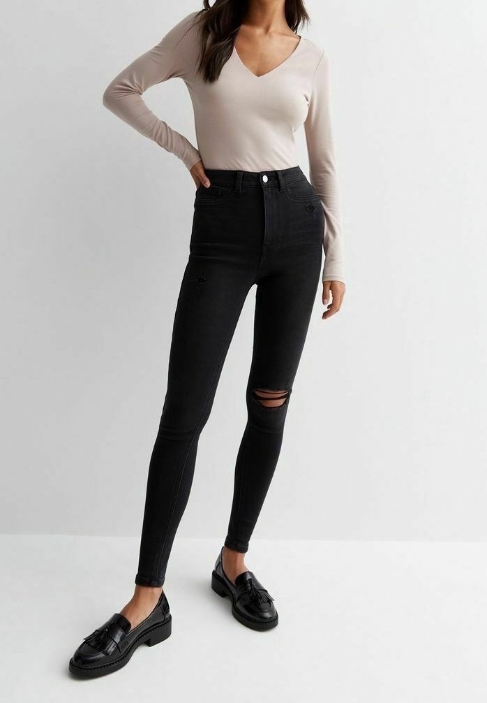 Jeans negros de cintura alta con corte ajustado, que presentan un desgaste en la rodilla, combinados con una blusa de manga larga en color beige claro con escote en V y mocasines negros.