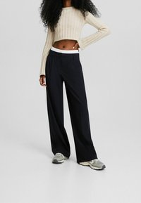 Kort beige ribbstickad tröja med långa ärmar, matchad med löst sittande svarta byxor med vit midja. Avslappnade grå sneakers.