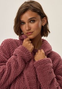 Puchowa, teksturowana różowa kurtka z kołnierzem i dużymi rękawami, o przytulnym designie odpowiednim do noszenia na cebulkę. Miękki materiał zwiększa ciepło.