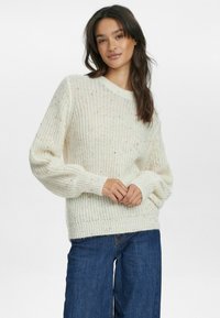 Cremfarbener Strickpullover mit lockerer Passform, geripptem Halsausschnitt und strukturiertem Muster mit bunten Sprenkeln. Kombiniert mit dunkelblauen Jeans.