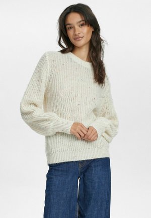 Crème-kleurige gebreide trui met een losse pasvorm, geribde halslijn en een getextureerd patroon met kleurrijke spikkels. Gecombineerd met donkerblauwe jeans.