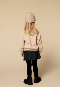 Kind met lang blond haar, gekleed in een beige gebreide muts, een beige trui, een zwarte rok, zwarte panty’s en zwarte laarzen, staand met de rug naar de kijker.