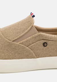 Beige canvas instapschoen met een gestructureerd oppervlak, elastische zijpanelen, een witte rubberen zool en een klein rood, wit en blauw label aan de hiel.