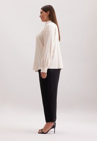 Polaire blouse in lichtcrème met lange mouwen en een hoge halslijn, gecombineerd met zwarte, op maat gemaakte broeken en zwarte, high-heeled sandalen.