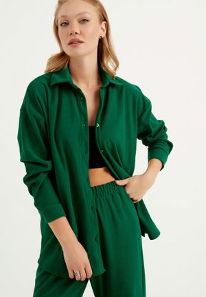 Chemisier vert à rayures avec des manches longues sur un top noir, assorti à un pantalon vert à taille élastique, mettant en avant un look décontracté.