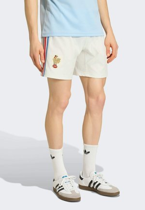 Person trägt weiße Shorts mit einem Hahn-Emblem, weiße Adidas-Socken und weiße Adidas-Sneaker mit schwarzen Streifen.