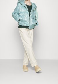 Person in einer hellblauen, gesteppten Parka-Jacke, dunkelgrünem Hemd, cremefarbener, weiter Hose und beigen, klobigen Stiefeln, der mit Händen in den Taschen dasteht.