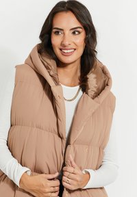 Gilet matelassé beige avec une grande capuche, doté d'une doublure brillante. Le tissu semble doux, avec des boutons-pression et une coupe décontractée. Accent doré visible.