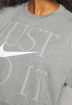 Osoba nosząca szarą koszulkę z okrągłym dekoltem z dużymi literami układającymi się w napis „JUST DO IT” oraz białym logo Nike Swoosh na piersi.