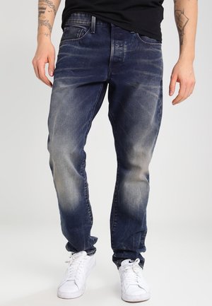 Abgetragene, dunkelblaue, ausgebleichte Jeans mit lockerem Schnitt, kombiniert mit weißen Sneakern und einem schwarzen Hemd, sichtbar tätowierte Arme.