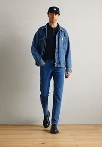 Denimjakke med to brystlommer, sort polo t-shirt, blå jeans og sorte sko; modellen har en sort cap med logo på.