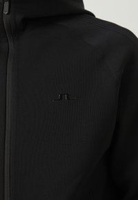 Svart hettegenser med glidelås laget av teksturert stoff med ribbestruktur. Har en liten, blank logo på brystet.