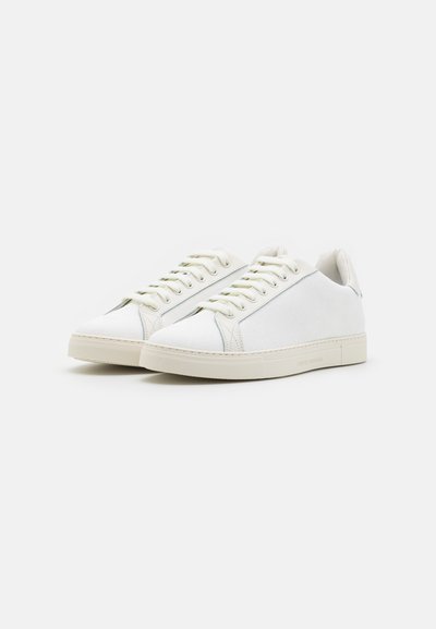 White sneakers made of textured material with flat laces, a round toe, and a flat rubber sole. Accents include light stitching details.