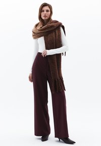 Bufanda marrón de gran tamaño, camiseta blanca de manga larga y pantalones de pierna ancha burdeos. Puntos de textura en la bufanda; tela suave. Zapatos de punta.