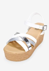 Sandales compensées blanches avec un design à brides croisées et des accents argentés, semelle en liège texturée et bride de cheville ajustable.