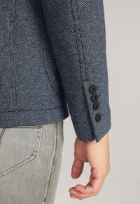 Textured dark blue blazer with a woven pattern; features four black buttons on the cuff; paired with light gray jeans.