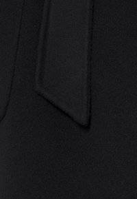 Tessuto di lana nero con superficie morbida e strutturata. Presenta un dettaglio prominente a fiocco piatto cucito sul materiale, che valorizza il design.