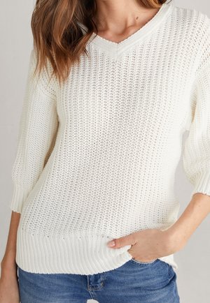 Vrouw draagt een wit gebreide V-hals trui met driekwart mouwen, hand in de zak, en een blauwe spijkerbroek, gefotografeerd van nek tot heupen.