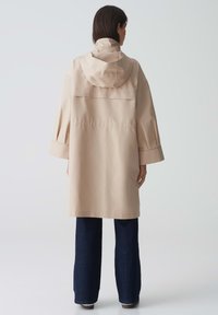 Giacca impermeabile beige con cappuccio, vestibilità ampia, maniche larghe e una ventilazione sul retro. Abbinata a pantaloni a gamba larga blu scuro.