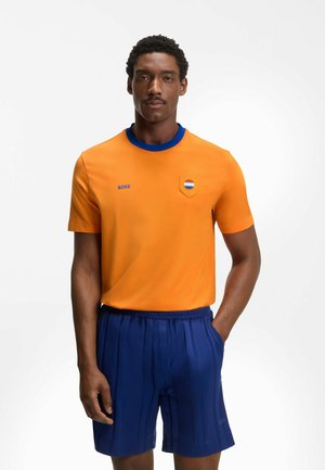 Man in een oranje T-shirt met korte mouwen en een blauwe kraag met logo, gecombineerd met een blauwe plissébroek, poseert met één hand in de zak.