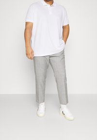 Mężczyzna w białej koszulce polo, szarych, w kratkę spodniach 3/4, białych skarpetach i białych trampkach z czarnym logo, stojący na tle jednolitym.