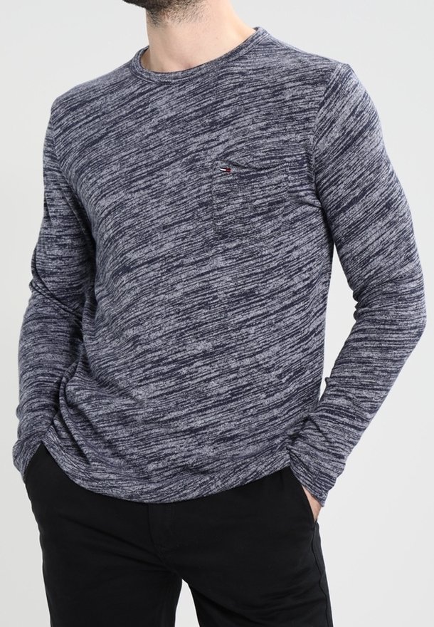 Homme portant un pull gris chiné à col rond et manches longues avec une petite poche poitrine, un pantalon noir, les mains dans les poches.