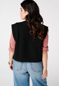 Zwarte textuurvest met een uitgesproken patroon, gedragen over een lichtroze kanten blouse. Gecombineerd met lichtblauwe, wijde denim jeans.