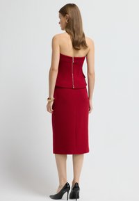 Rotes trägerloses Kleid mit einem taillierten Oberteil und Bleistiftrock, ausgestattet mit einem sichtbaren Reißverschluss auf der Rückseite. Getragen mit schwarzen hohen Schuhen.