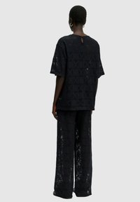 Schwarzes Zweiteiler-Outfit mit einer lockeren, strukturierten Bluse mit geometrischen Mustern und weiten Hosen, beide mit einer durchsichtigen Qualität und einem Schlüsselloch-Design im Rücken.
