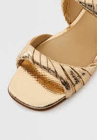 Χρυσό σανδάλι με υφή και τακούνι, με πλεγμένους ιμάντες, τετράγωνη μύτη και λεία εσωτερική σόλα. Η εξωτερική επιφάνεια έχει λαμπερό, αντανακλαστικό φινίρισμα.