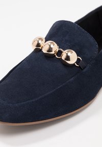 Mocasín de ante azul marino con un diseño plano, acentos decorativos redondos en tono dorado y una textura suave en la parte superior y la suela.