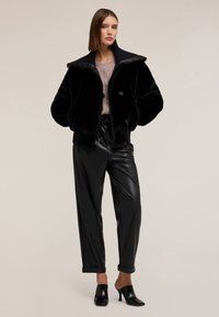 Giacca in pelliccia sintetica nera con ampio colletto e chiusura a bottone, abbinata a pantaloni in pelle sintetica neri e sabot con tacco, su uno sfondo neutro.