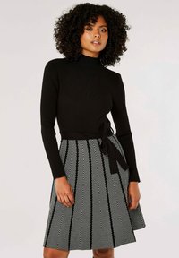 Haut noir à manches longues côtelé associé à une jupe à motif chevrons noir et blanc, dotée d'une taille nouée. Silhouette évasée, texture lisse.