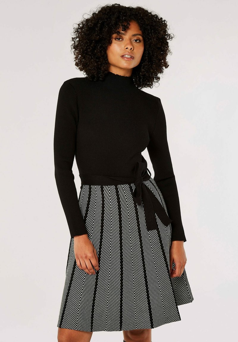 Haut noir à manches longues côtelé associé à une jupe à motif chevrons noir et blanc, dotée d'une taille nouée. Silhouette évasée, texture lisse.