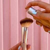 Rund makeupborste med mjuka, gradientborst, silverfärgad ferrule och ett beige handtag, som tar emot flytande foundation från en glasflaska.